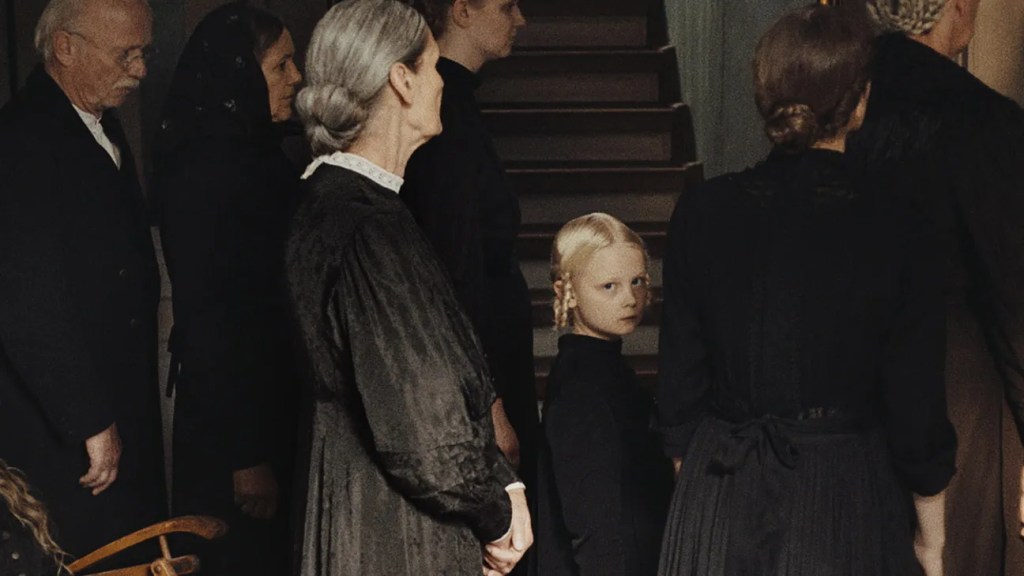 A young girl looks back hauntingly into the camera among a group of older people dressed smartly in black