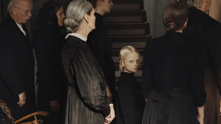 A young girl looks back hauntingly into the camera among a group of older people dressed smartly in black