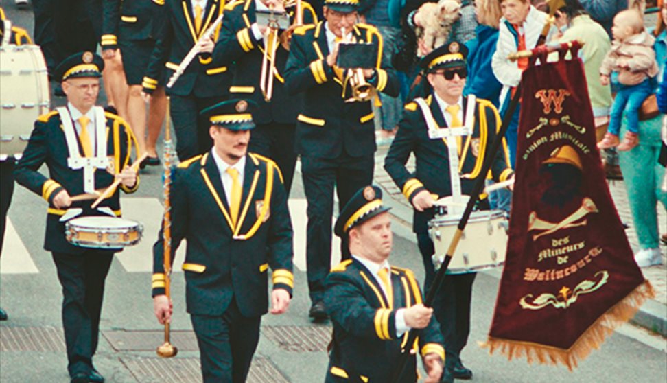 A marching band play instruments while parading down a street with flags, dressed smartly in suits.