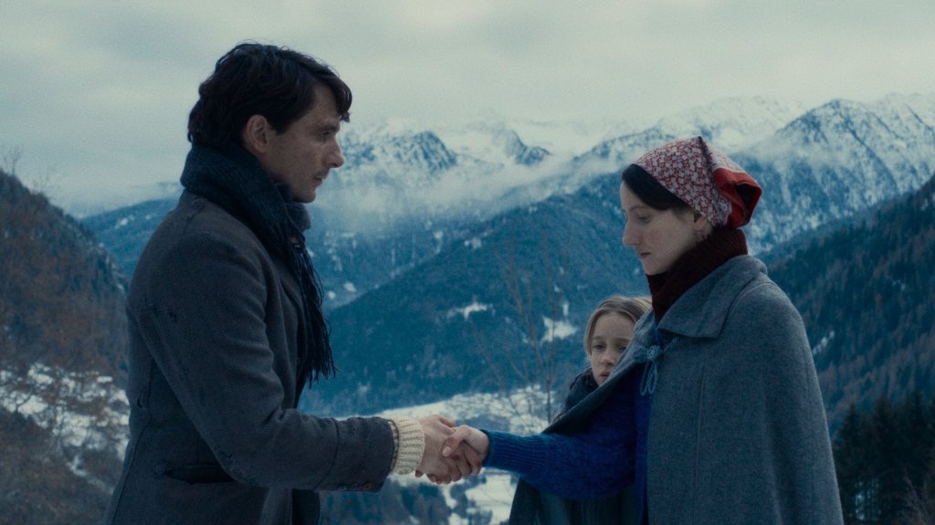 A man and woman shake hands while a young girl watches on against the backdrop of the snowy Italian alps