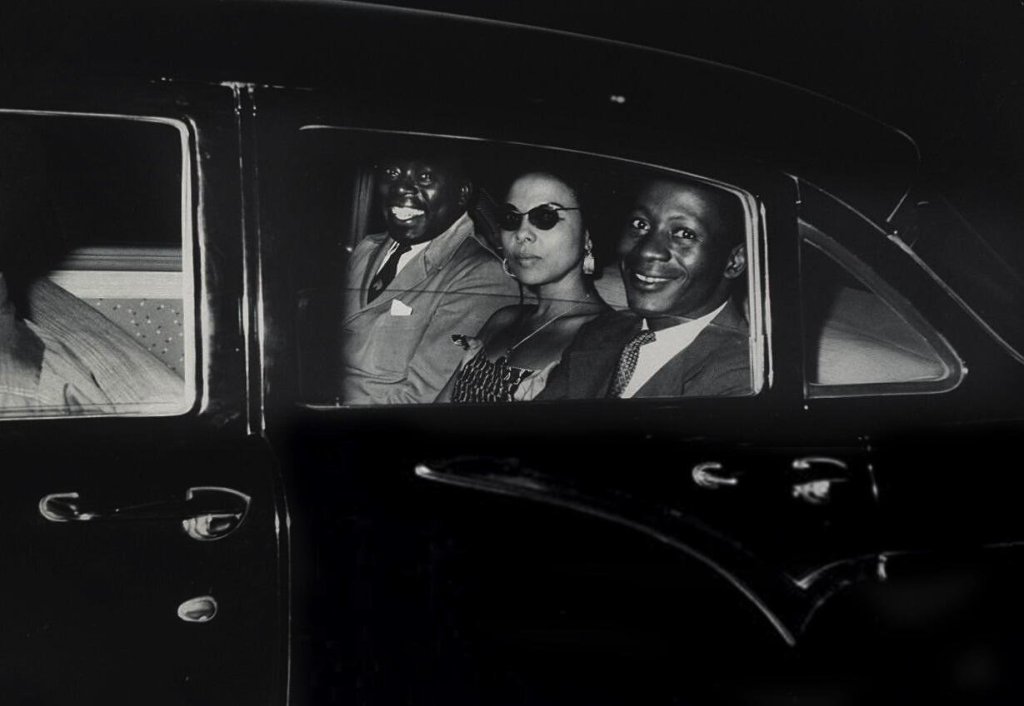 Black and white photo of two smiling suited men sat in the back of a car with a woman between them